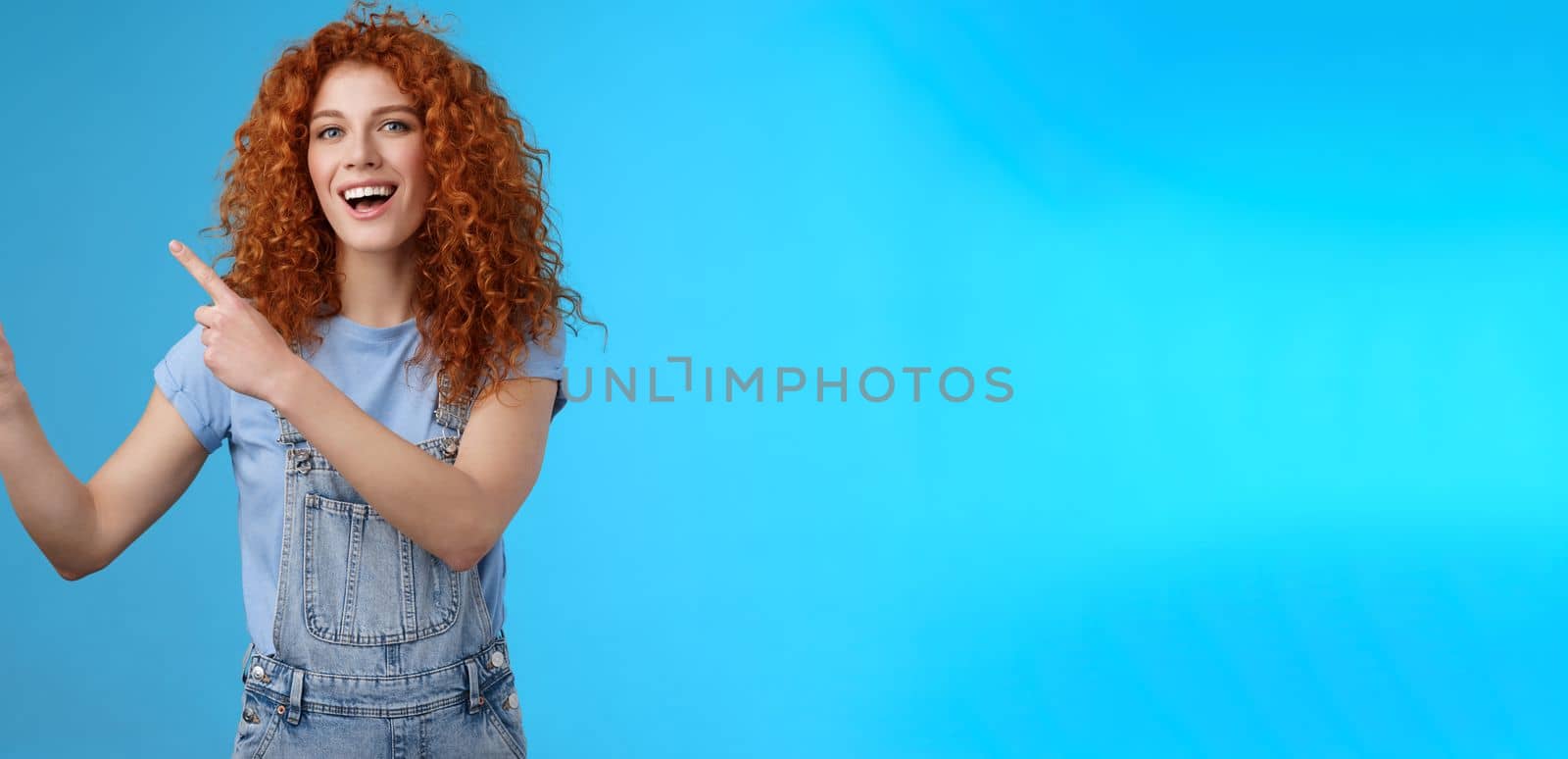 Hey check this out. Enthusiastic redhead curly stylish summer girl inviting beach party pointing upper left corner directing awesome location smiling broadly discuss awesome promo blue background by Benzoix