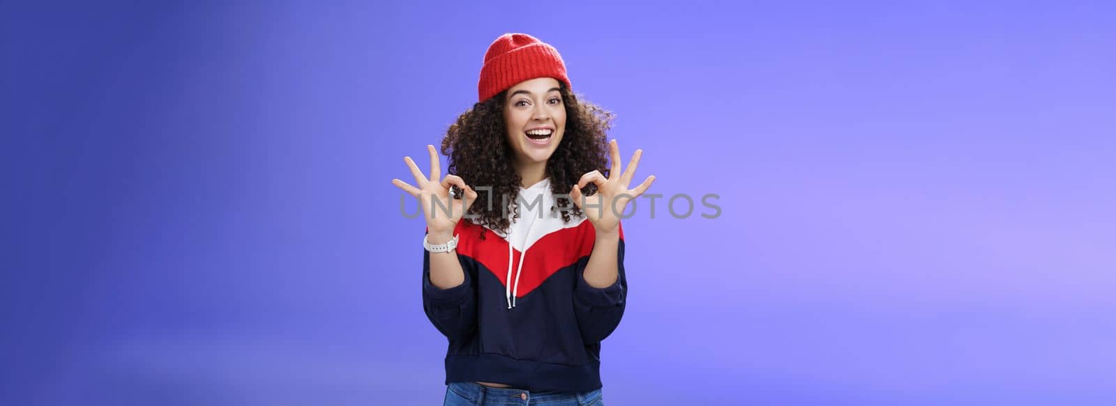 Got under control. Portrait of happy charming smiling curly-haired female in warm winter hat and sweatshirt smiling broadly and showing okay or excellent gesture as approving, liking cool movements by Benzoix