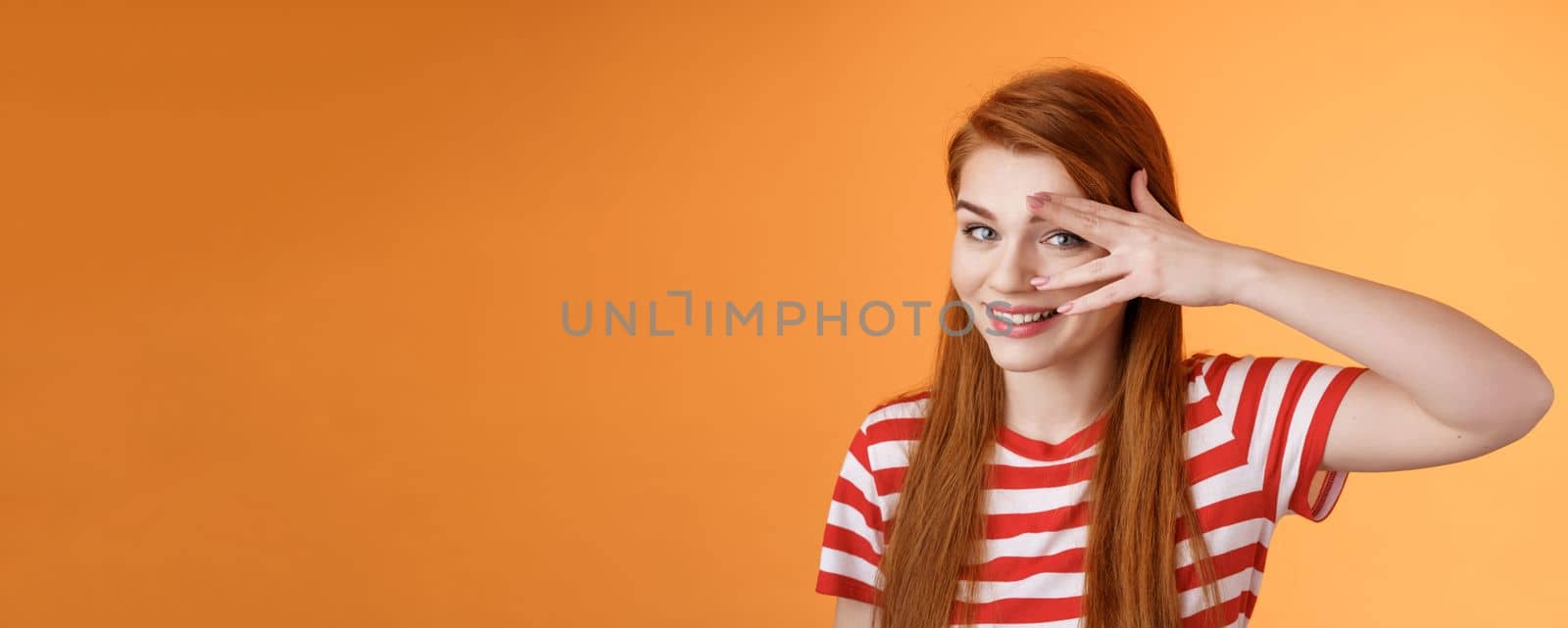 Charismatic lovely redhead lucky female, smiling cute, peek camera tender coquettish gaze, hold palm face, look through fingers, stand orange background optimistic, cheerful mood. Copy space