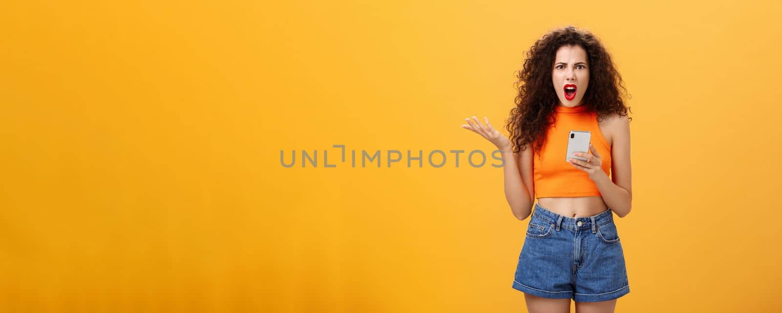 Portrait of displeased pissed and irritated caucasian female with curly hairstyle in red lipstick and orange cropped top holding smartphone and gesturing with palm annoyed complaining on dumb message by Benzoix