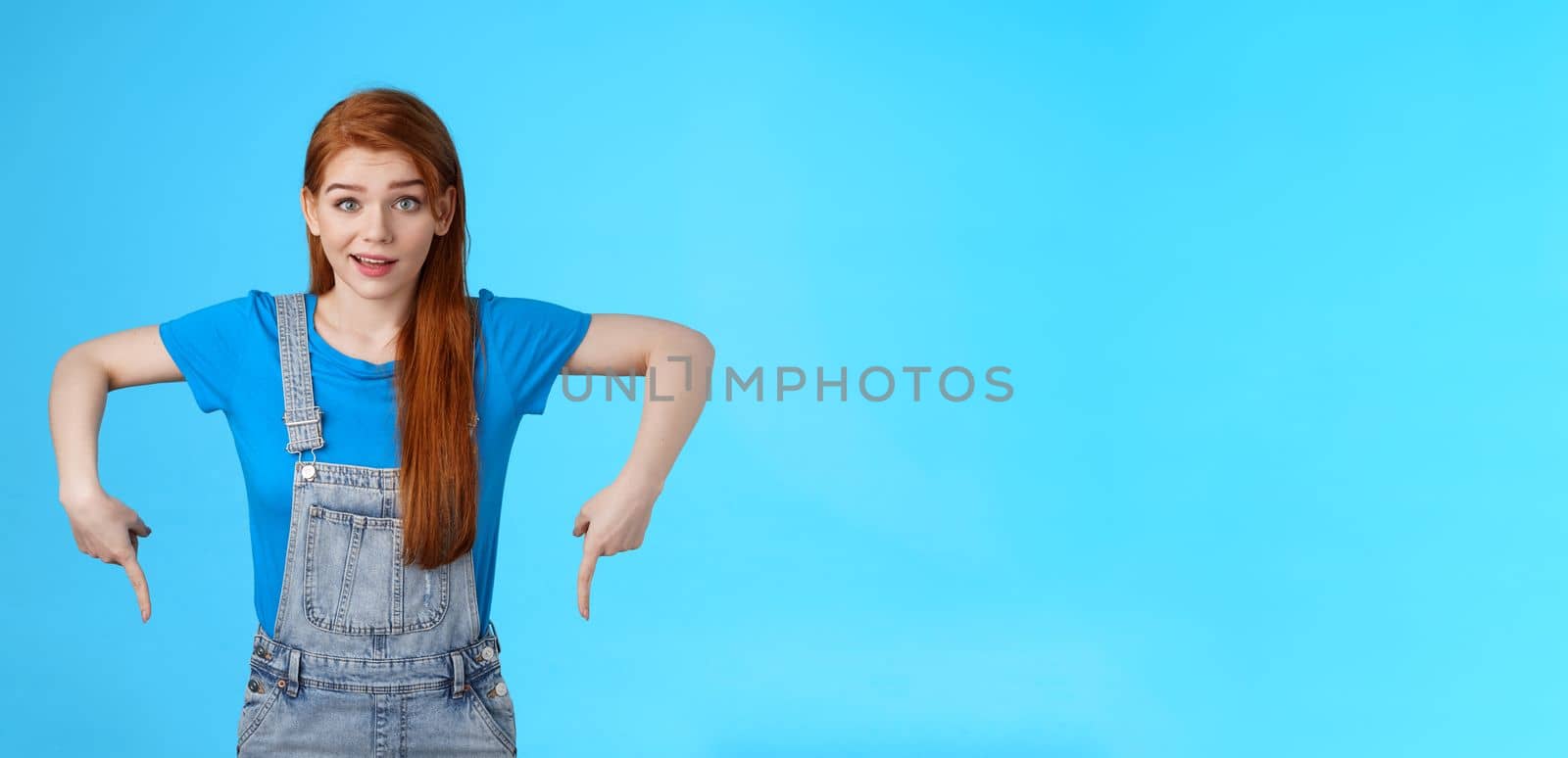 Curious tender lovely ginger caucasian girl, pointing down questioned, check out interesting promo, point bottom down copy space, look camera intrigued, suggest good product online shop link.