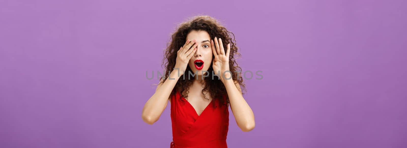 Wow awesome take look. Amazed and surprise curious caucasian woman with curly hair in red stylish dress open mouth from amazement and joy peeking with one eye while closing sight with palms.