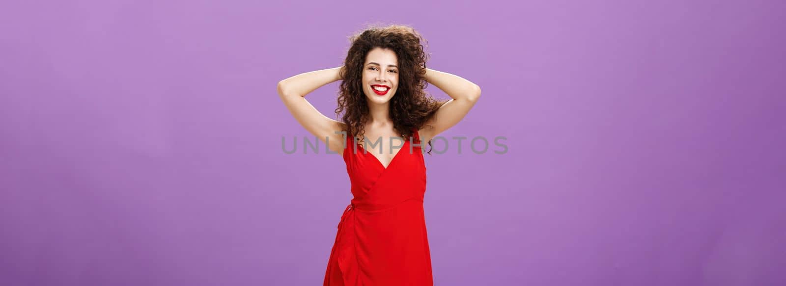 Feeling alive and energized like queen of show. Carefree elegant curly-haired woman in stylish red evening dress playing with hair and smiling broadly feeling beautiful in new outfit over purple wall.