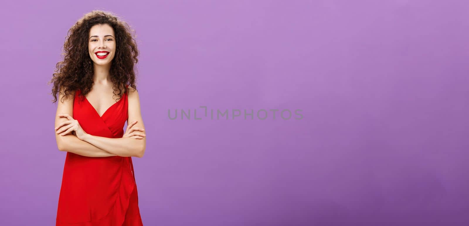 Cute ambitious and happy good-looking european woman. in luxurious red dress holding hands crossed in self-assured gesture smiling broadly having party celebrating graduation over purple background.