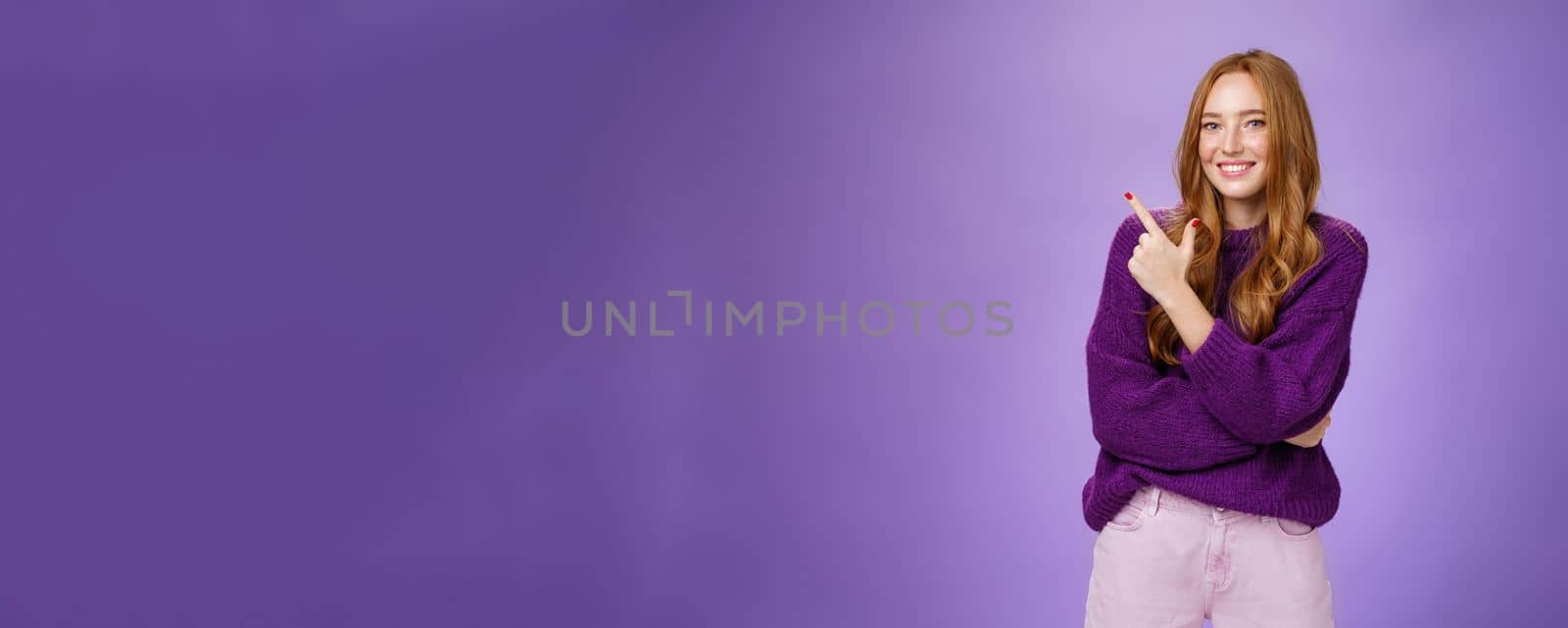 Lifestyle. Charming friendly-looking bright redhead woman with freckles and makeup in purple warm sweater pointing at upper left corner and smiling delighted and cute at camera as showing cool place hang out.
