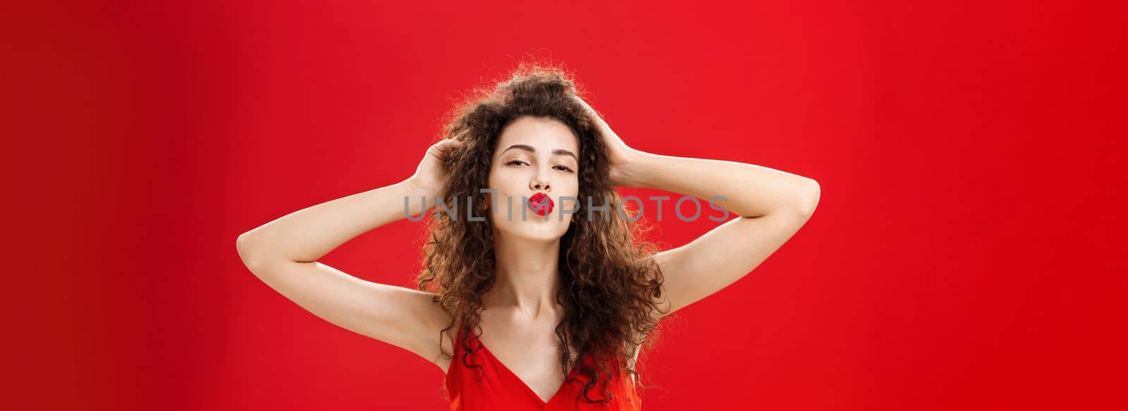 Waist-up shot of confident flirty and elegant young rich woman. with curly hairstyle in red lipstic and evening dress touching hair folding lips in mwah giving kiss to admirer feeling self-assured.