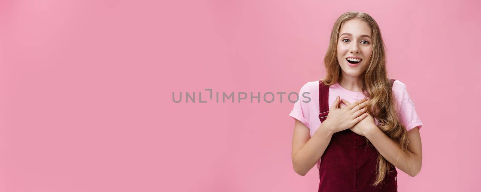 Portrait of thankful and pleased happy young girl receiving surprise gift on b-day pressing palms to hear in gratefyl gesture open mouth from amazement and joy thanking over pink background by Benzoix