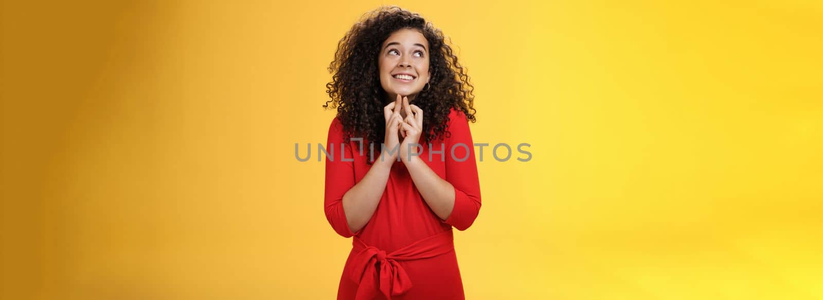 Hopeful, excited cute tender woman in red dress with curly hair standing new x-mass tree crossing fingers for good luck and smiling with head raised to sky making wish, having faith in dream come true by Benzoix