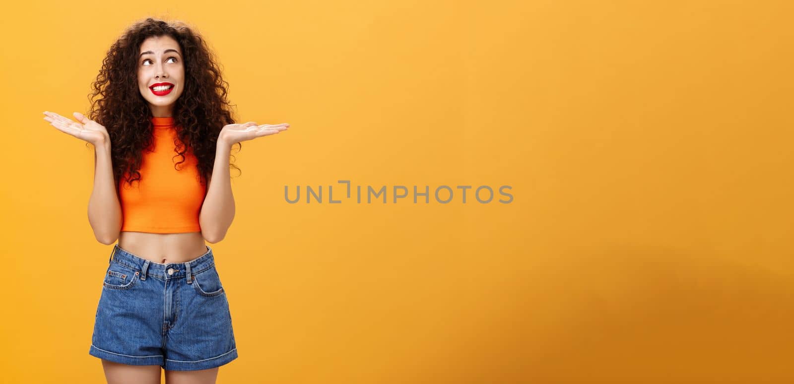 Silly and clueless carefree cute girl. with curly hairstyle in cropped top and shorts shrugging with palms spread aside smiling unaware and gazing at upper right corner uncertain and uninvolved.