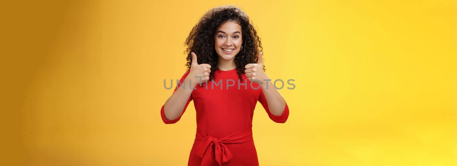 Excellent idea, nice job. Portrait of supportive delighted and happy charming female friend showing thumbs up as standing in red dress over yellow wall smiling giving positive reply, liking concept by Benzoix