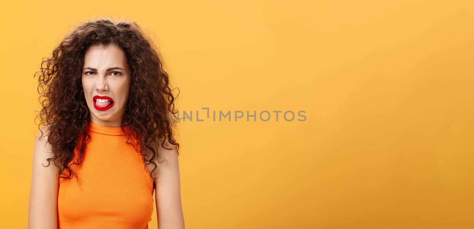 Girl being disgusted with row food sticking out tongue and frowning showing aversion and disgust talking about thing she dislikes with friend standing intense and dissatisfied over orange background. Copy space
