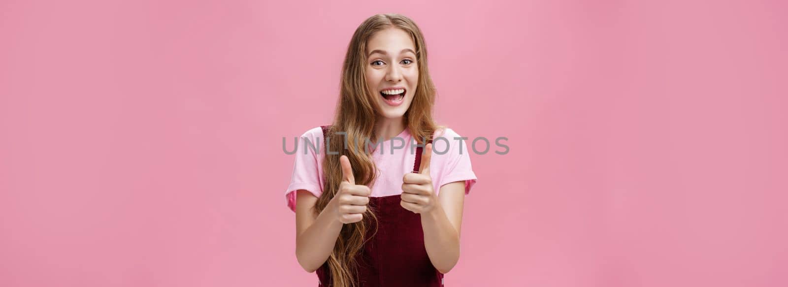 Friendly and outgoing pretty young woman in overalls with wavy natural hairstyle showing thumbs up gesture with cheer and joy smiling being supportive, liking idea, expressing positive opinion by Benzoix
