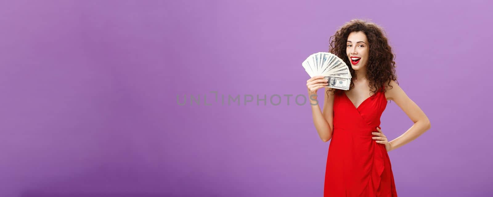 Girl knows how earn money. Successful and delighted satisfied woman in elegant red dress with curly hair holding cash near face like hand fan rejoicing from lots of cash in arms, smiling pleased.