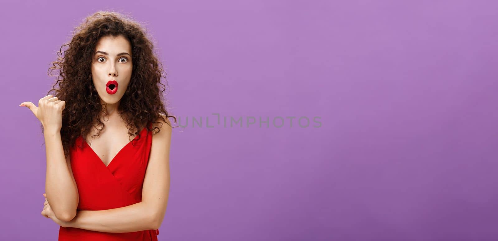 Curious and questioned amazed woman. with curly hairstyle in stylish red dress folding lips in wow sound pointing left with thumb asking question about stunning and thrilling scene over purple wall.