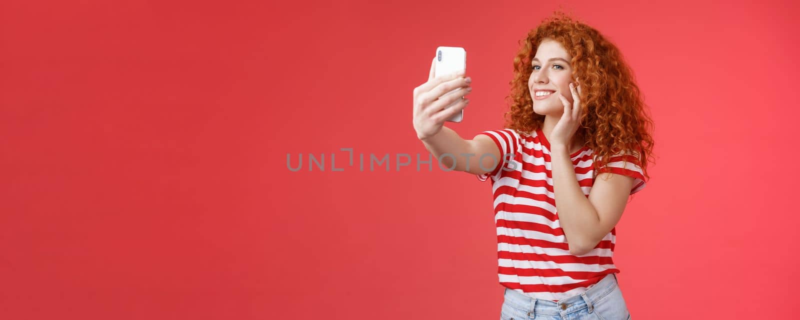 Popular cheerful good-looking stylish female blogger redhead curly hairstyle feel pretty self-acceptance taking selfie raised arm holding smartphone posing silly cute phone camera red background by Benzoix