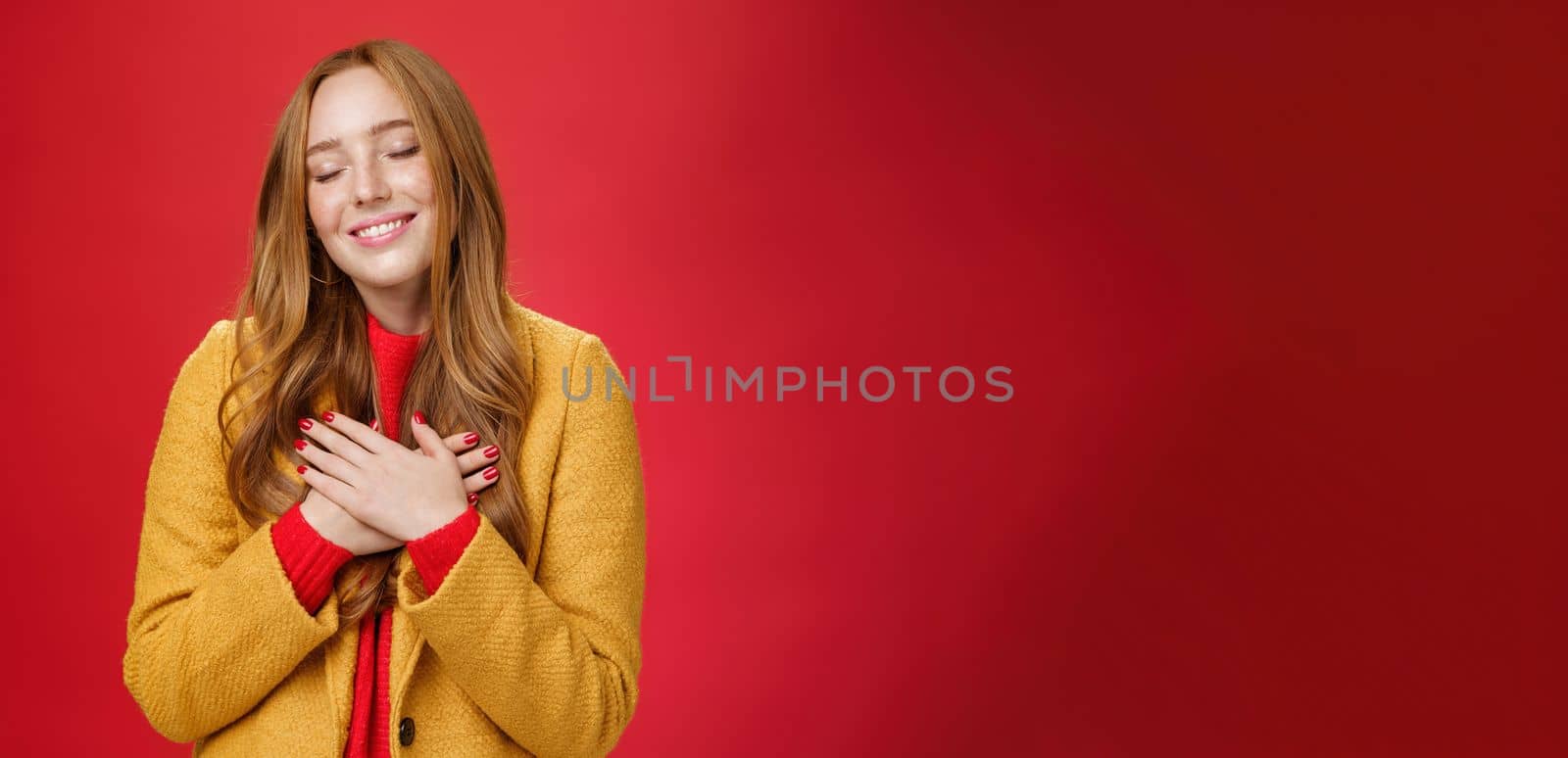 Girl feeling like heart warming as remembering sweet tender memories with closed eyes and silly broad smile holding hands on chest pressing to soul posing nostalgic and touched over red background by Benzoix