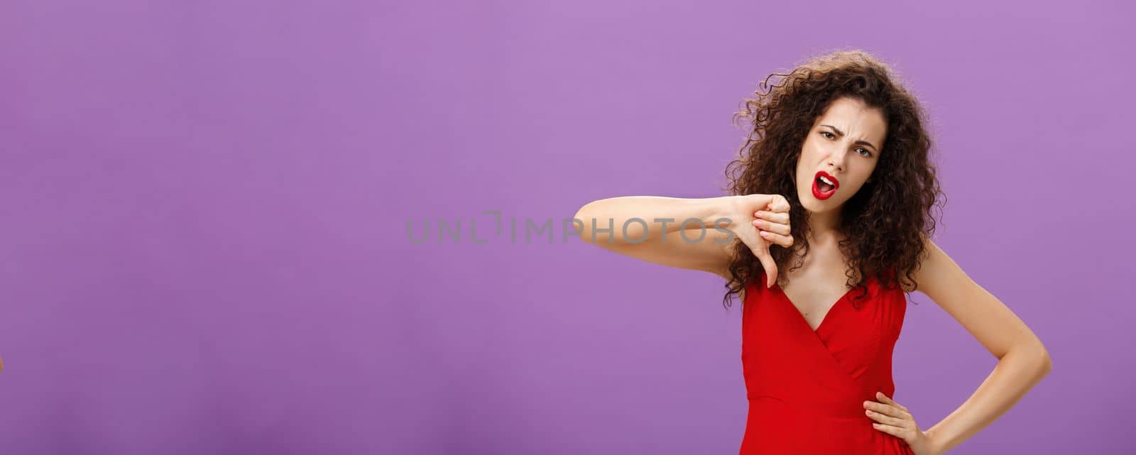 Unimpressed arrogant and picky stylish rich woman with curly hairstyle and evening make-up holding hand on waist stooping shoulders showing thumbs down and grimacing from dislike, being dissatisfied.