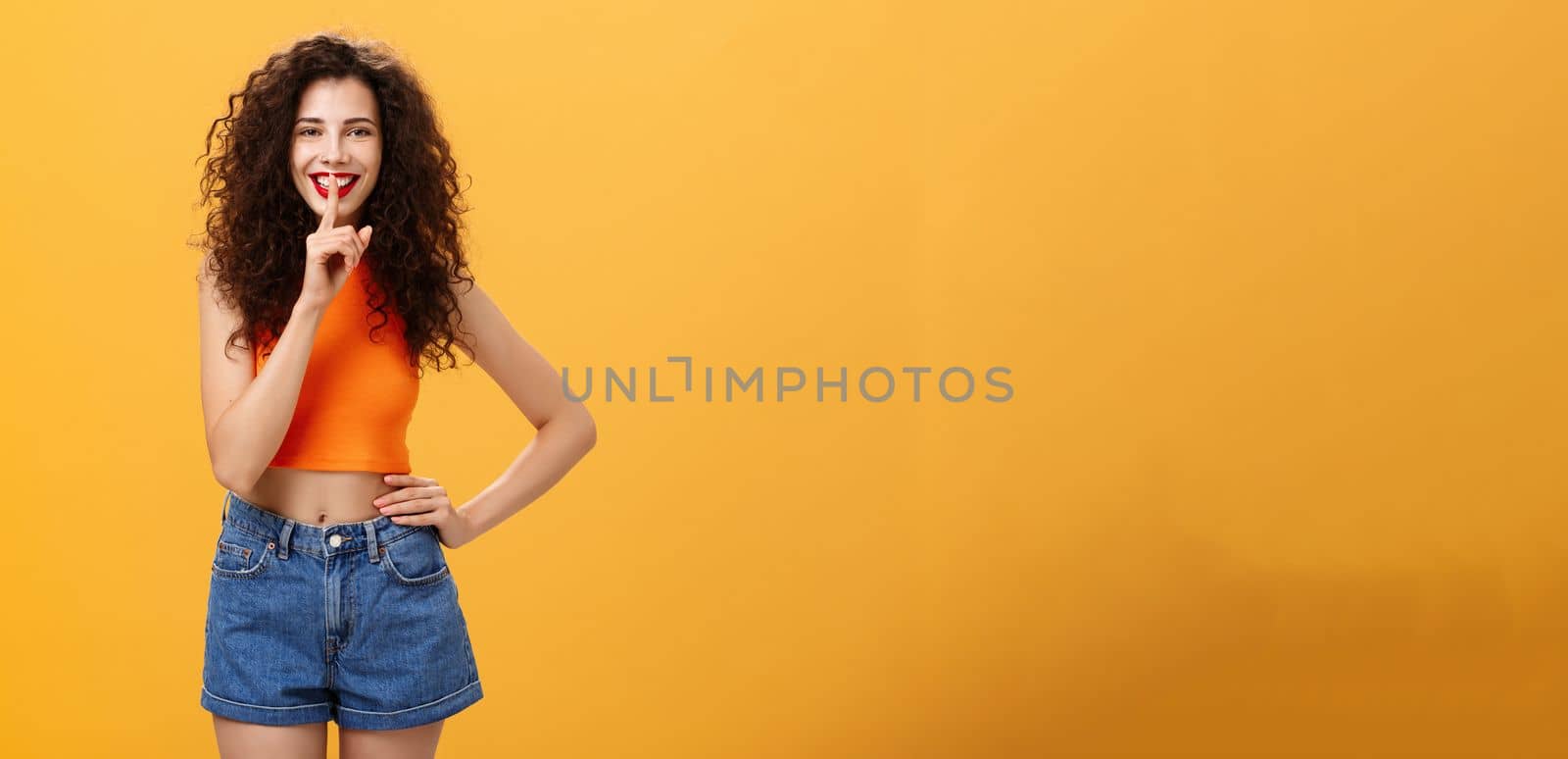 Shh I got something for you. Portrait of charming european young female with curly hairstyle in cropped top and shorts showing shush gesture with broad smile having secret preparing surprise.