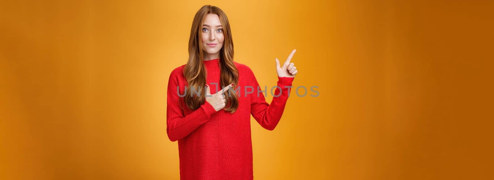 Soft and gentle gorgeous ginger girl with freckles in red warm sweater smiling joyfully pointing at upper left corner smiling showing curious place, suggesting visit it over orange background.