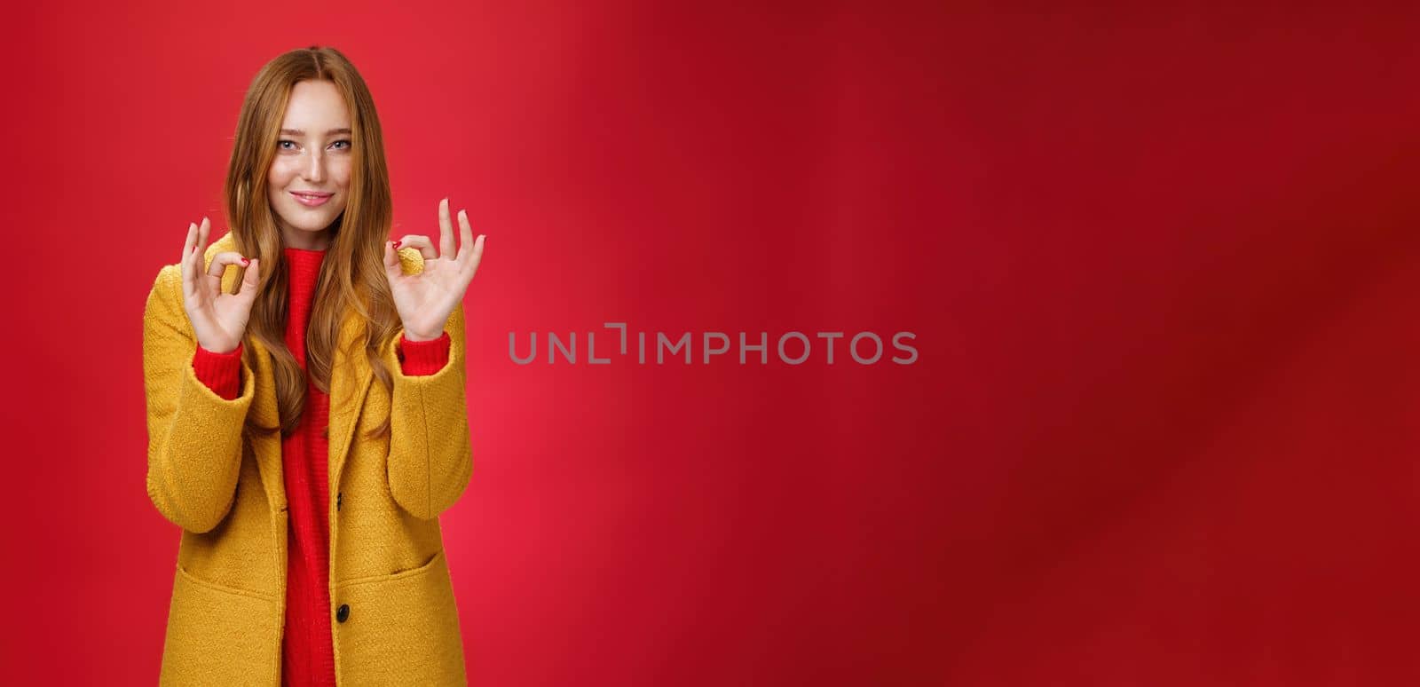 Indoor shot of pleased bright and happy young woman feeling okay and perfect in warm comfortable yellow coat picking right close for cold weather showing ok gesture and smiling delighted over red wall. Fashion and temprature concept