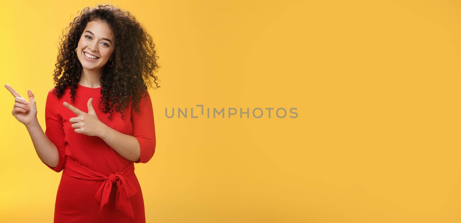 Friendly cute assertive female assistant in elegant red dress with curly hair tilting head flirty and smiling broadly, satisfied as pointing at upper left corner, recommending product over yellow wall.