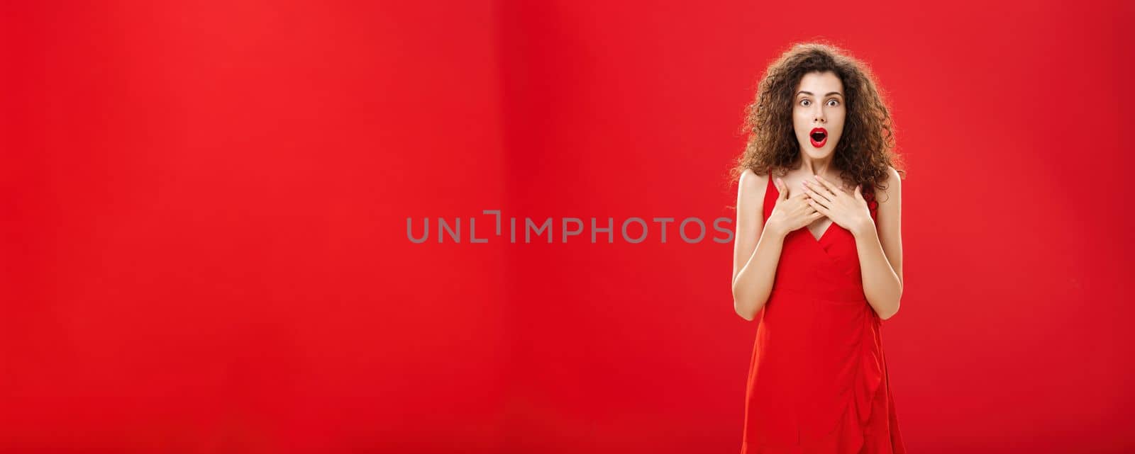 Woman receiving awesome news being impressed and amazed in red evening dress gasping saying wow holding hands on brest standing astonished and excited over red background gossiping. Copy space