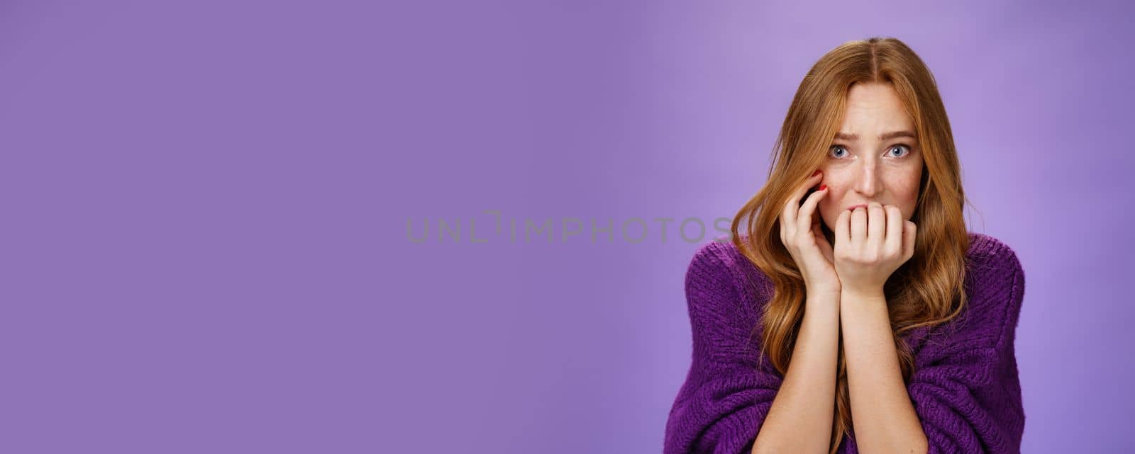 Scared insecure woman with red hair biting fingernails feeling frightened as home alone hearing weird horrifying noises shaking from fear standing anxious and worried over purple background by Benzoix