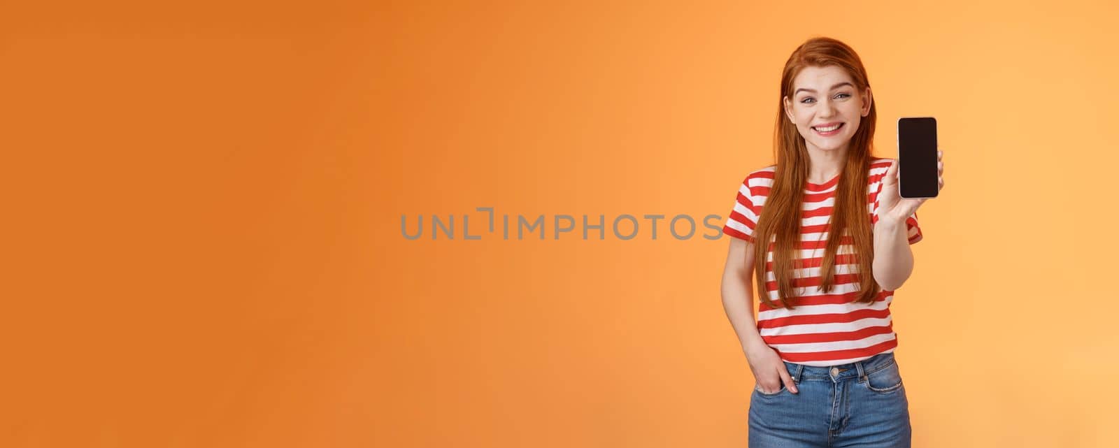 Cheerful good-looking 20s redhead woman introduce social media feature product hold smartphone, show camera cellphone display, promote device application, smiling broadly, orange background.