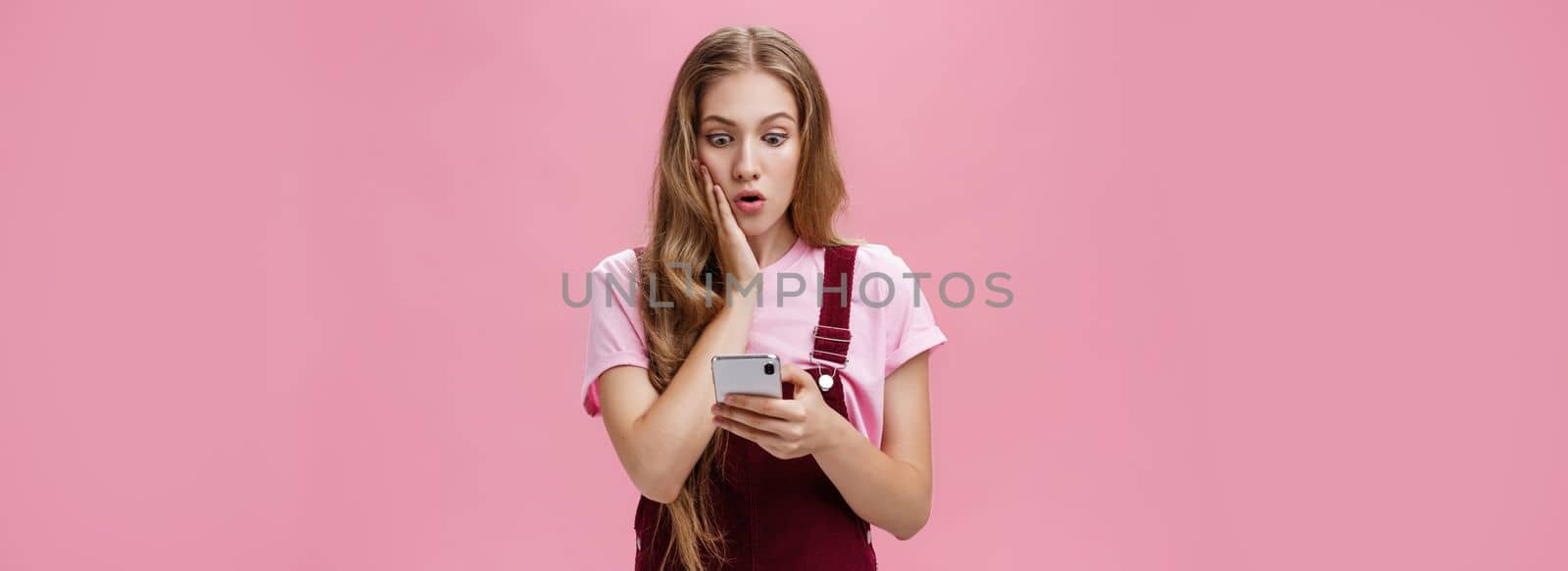 Girl found out shocking truth after reading message in smartphone pressing arm to cheek in surprise staring concerned and speechless at cellphone screen reacting to unexpected news over pink wall. Technology, emotions concept