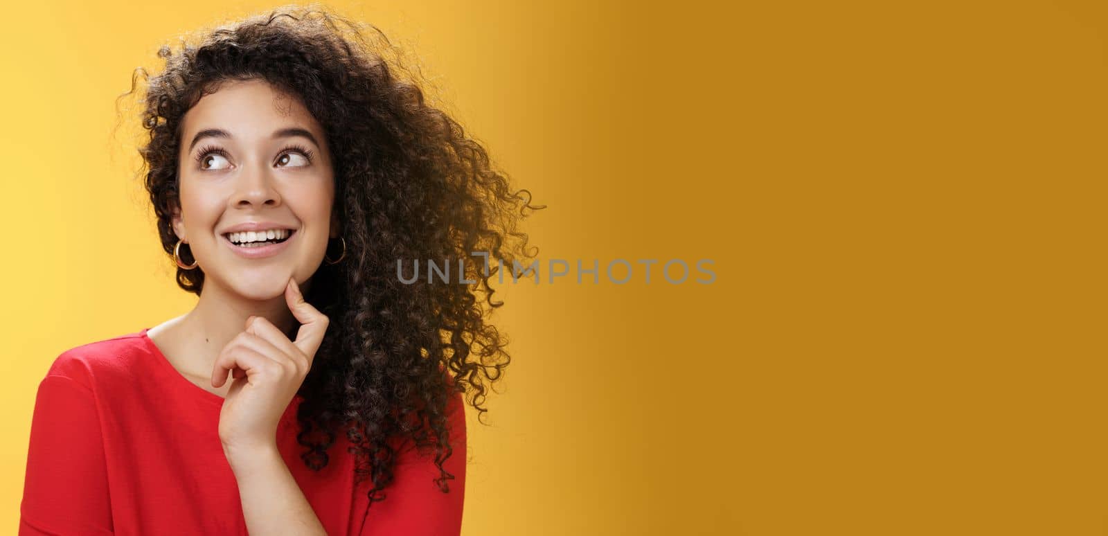 Dreamy girl making assumptions what gets as christmas present looking curious and happy at upper left corner, thinking, touching chin, standing thoughtful with desire and wish in mind over yellow wall.