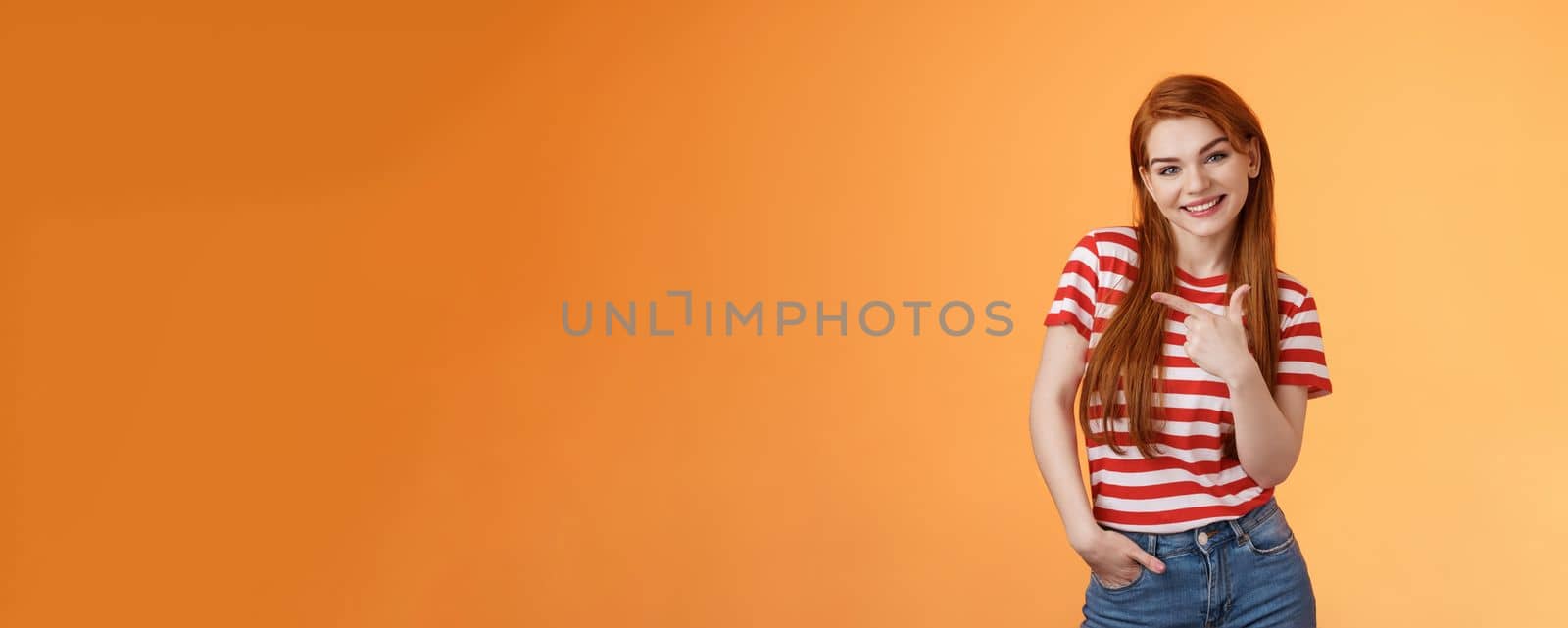 Tender cute feminine redhead lovely girl, tilt head cheerfully smiling, pointing left, showing way, give advice what purchase, advertising good product, introduce shop, orange background.