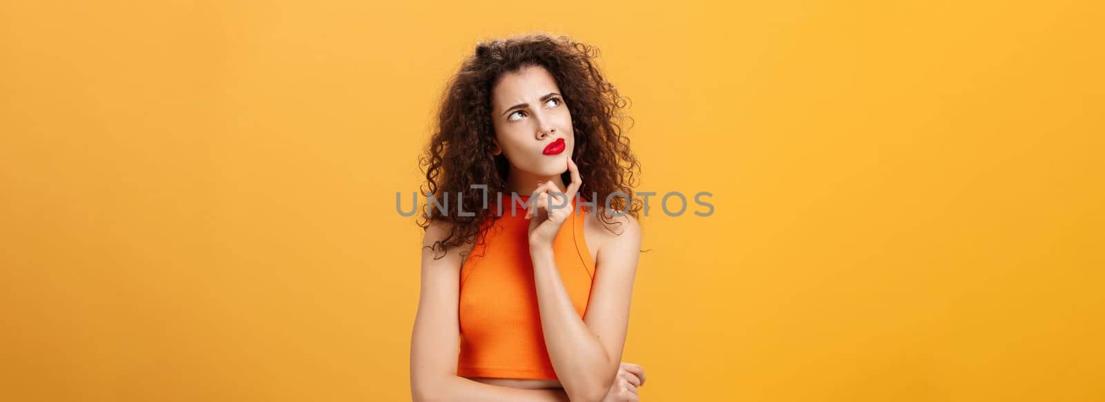 Waist-up shot of perplexed thoughtful. and smart female with curly hairstyle in red lipstick and cropped top holding in hmm pose touching chin frowning looking at upper left corner thinking determined.