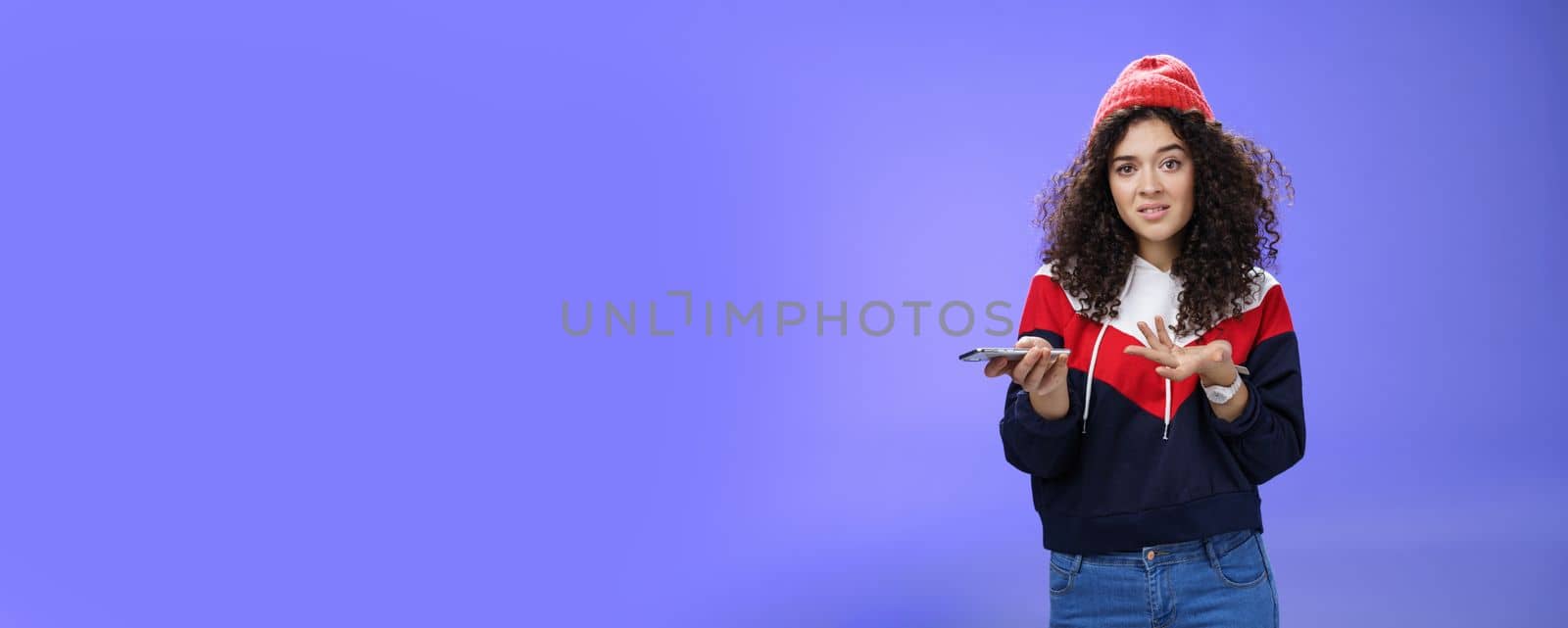 Confused and clueless girl cannot understand meaning of strange message shrugging grimacing questioned as holding smartphone and extending arm in unaware gesture over blue background by Benzoix