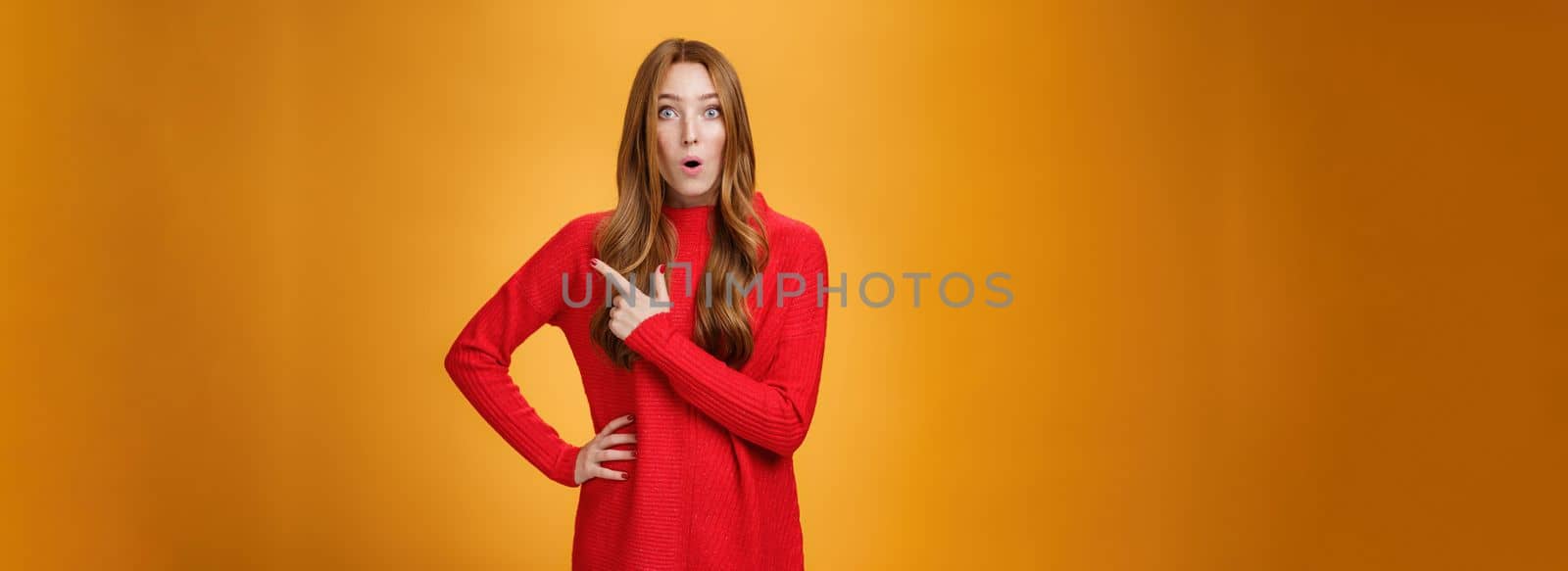 Impressed and surprised european ginger girl in red warm sweater open mouth from excitement and amazement pointing at upper right corner, gasping astonished and curious, asking question.