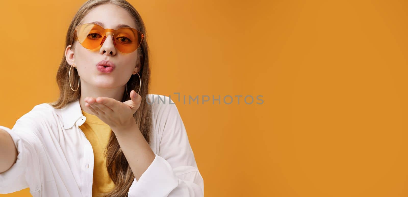 Stylish popular girl sending air kiss at camera taking selfie or recording vlog working as freelance travel advisor taking pictures against orange background tender and feminine pulling hand forward by Benzoix