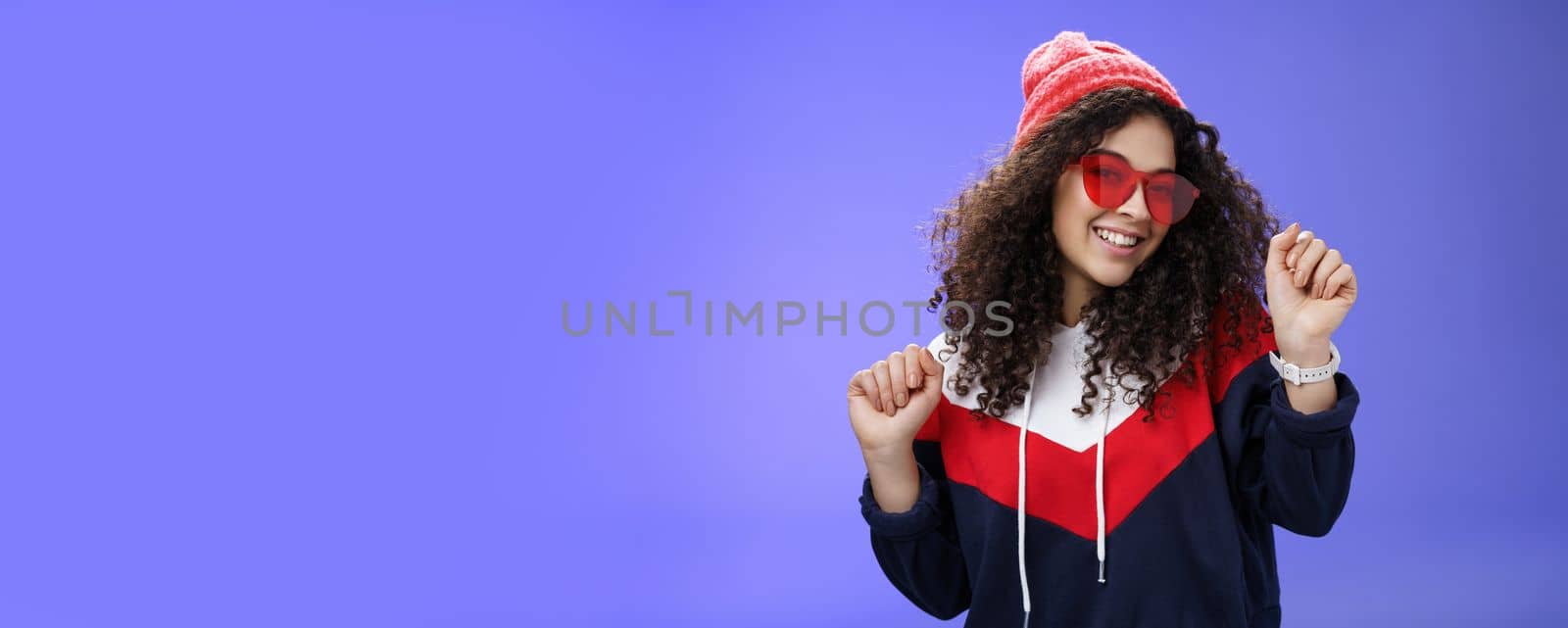 Stylish and feminine girlfriend dancing having fun feeling upbeat as liking weather feeling warm and cozy in stylish red beanie and sunglasses dancing with raised hands and broad smile over blue wall by Benzoix