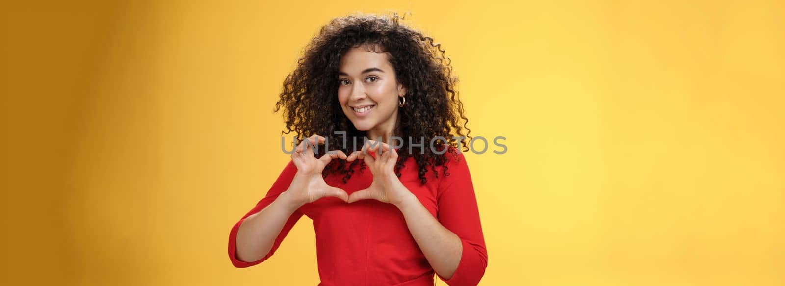 Waist-up shot of romantic and cute pretty girlfriend with curly hair in red dress showing heart sign over chest and smiling broadly confessing in admiration and love posing over yellow background by Benzoix