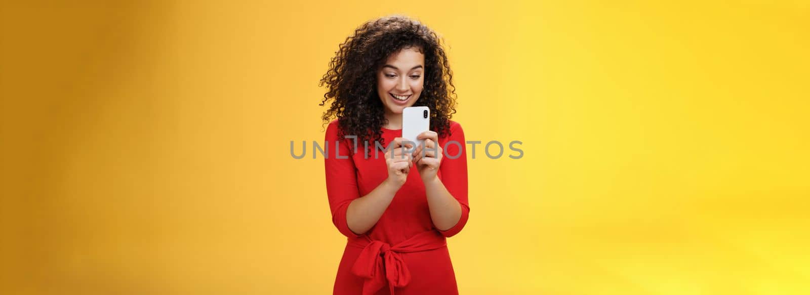 Wow new mobile phone amazing. Impressed and astonished good-looking curly-haired female in red dress holding smartphone looking at screen amused as playing cool app or game over yellow wall by Benzoix