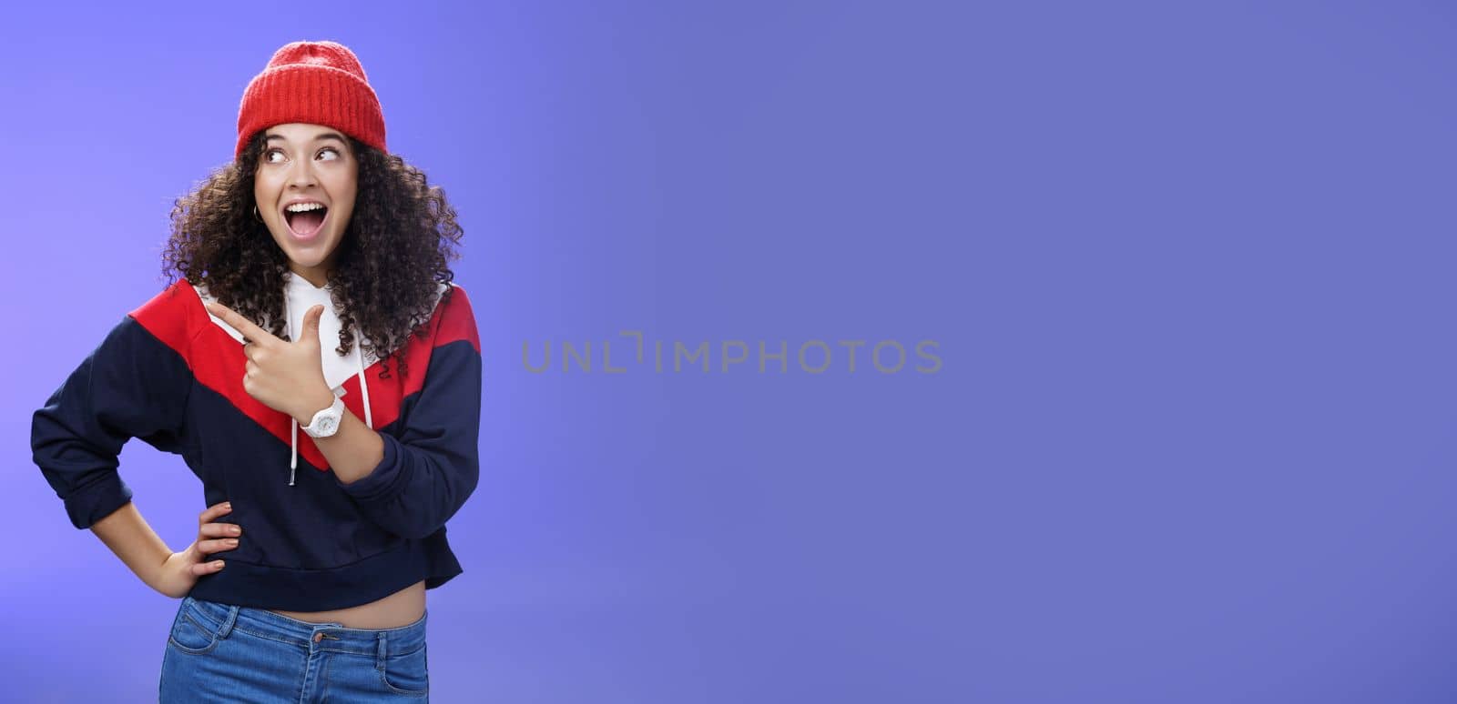 Cute and playful girl in winter stylish outfit and watch smiling broadly with excitement and joy pointing, looking at upper left corner thrilled and joyful as posing over blue background.
