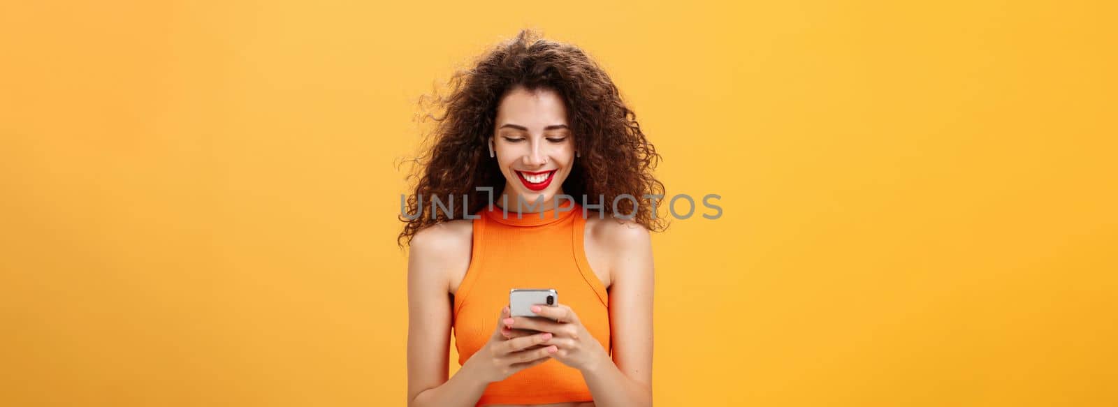 Stylish young attractive woman with curly hairstyle, nose ring and red lipstick picking song, typing in smartphone listening music in wireless earphones smiling at device screen over orange wall.