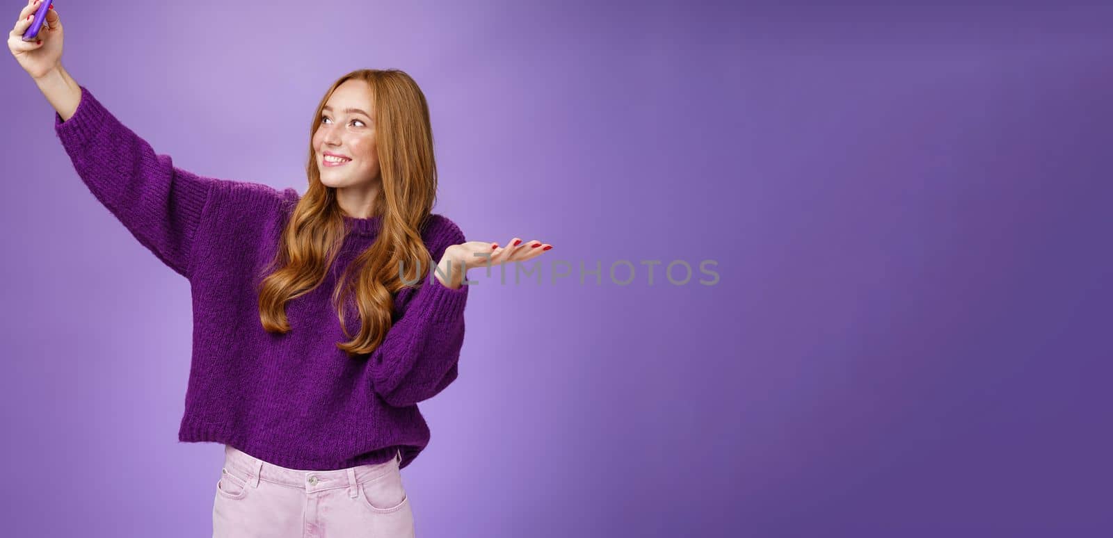 Portrait of cute redhead girl on vacation taking selfies near sightseeing raising hand as if holding builting on palm or showing cool place as taking photograph with pulled arm and smartphone by Benzoix