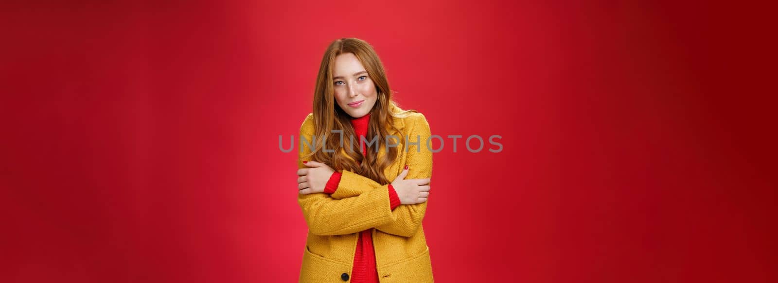 Romantic good-looking tender and cute ginger girlfriend. with freckles hugging herself and looking with sensual gentle smile at camera getting cold or chilly waiting outside over red background.
