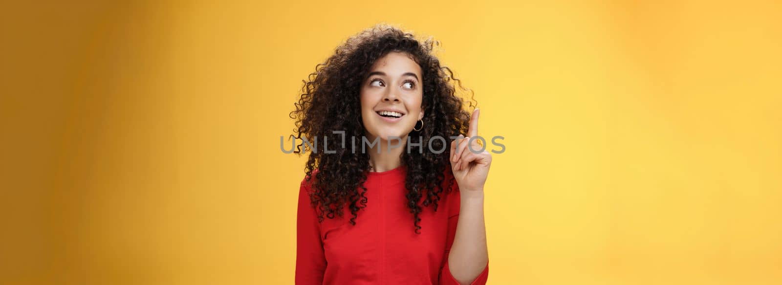 I got idea. Excited happy attractive woman with curly hair in red dress raising index finger in eureka gesture smiling amused, looking at upper left corner, sharing plan or suggestion over yellow wall by Benzoix