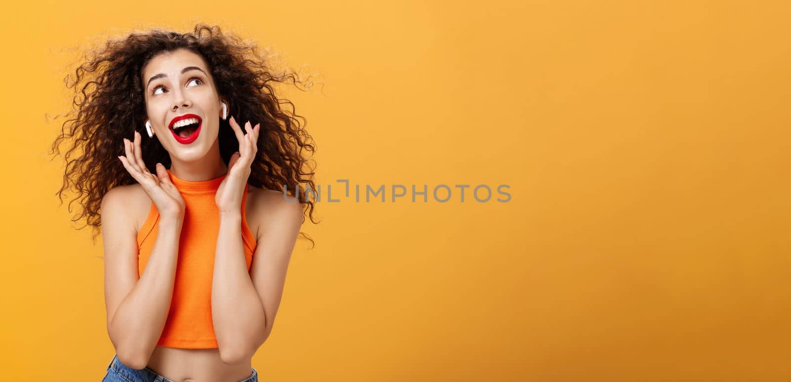 Waist-up shot of attractive silly curly-haired european female. in cropped top using wireless earphones touching earbuds and gazing at upper right corner delighted and carefree over orange wall.