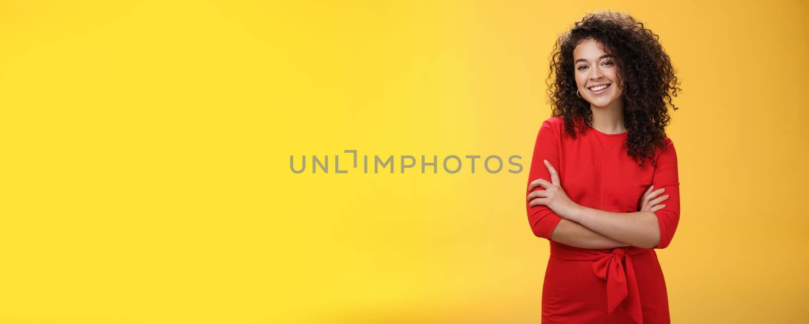 Confident charismatic young curly-haired woman in casual red dress holding hands crossed over chest in self-assured satisfied expression, smiling delighted posing over yellow background.
