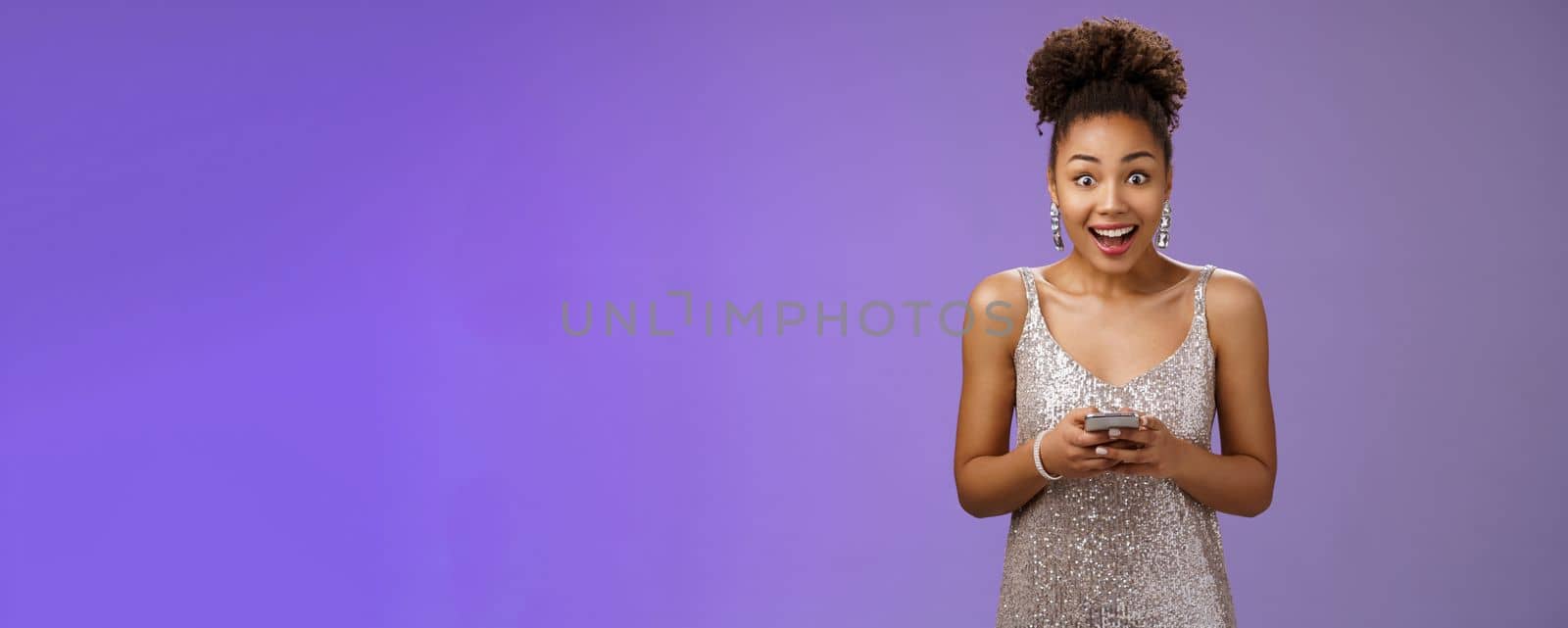 Amused african-american girl in silver shiny dress receive awesome surprising news via text message widen eyes impressed happily smiling unbelieveble luck standing amazed hold smartphone by Benzoix