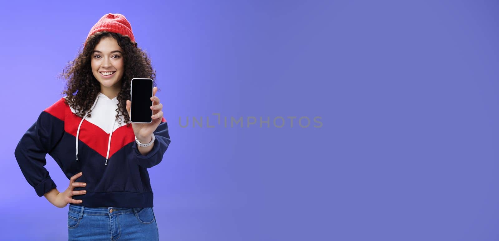 Girl showing her post as pulling smartphone screen towards camera smiling delighted waiting opinion, holding hand on waist in confident pose wearing warm winter red beanie and sweatshirt. Technology and people lifestyle concept