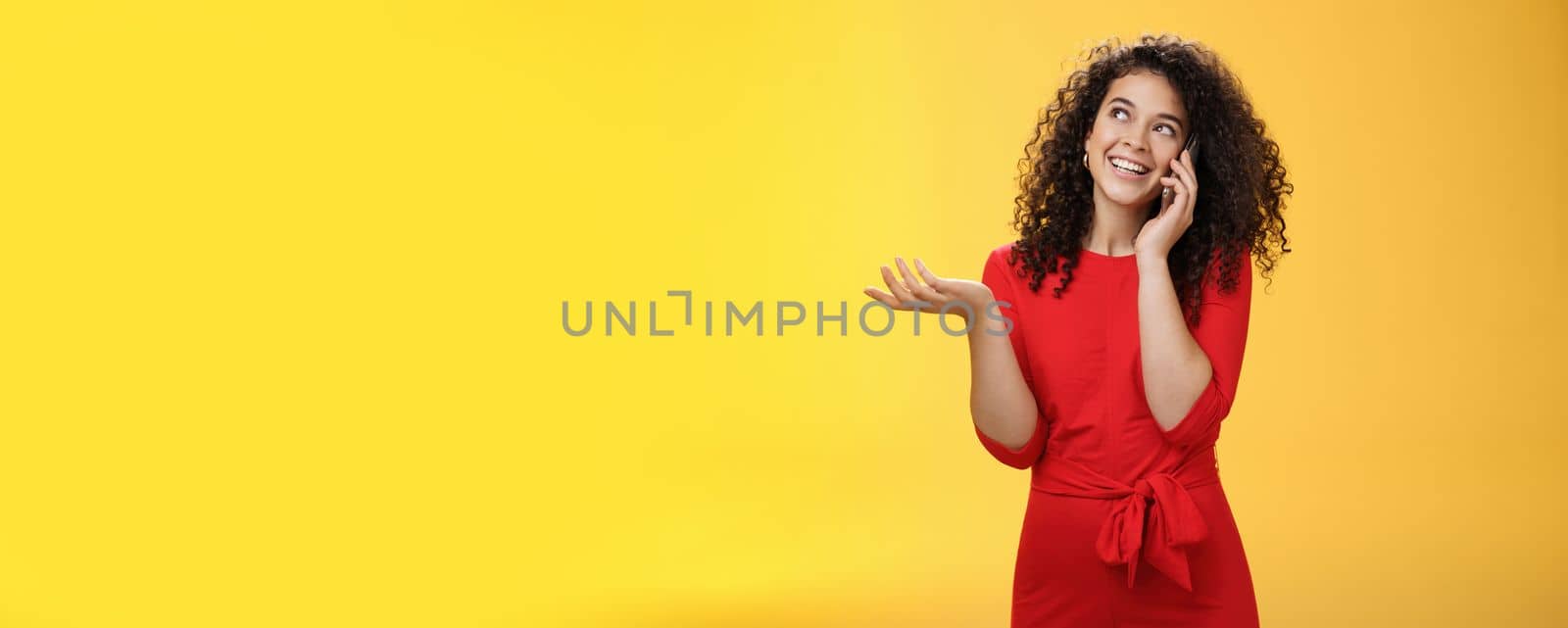 Carefree talkative cute woman with curly hair in red dress having char with friend on mobile phone holding smartphone near ear, gesturing as discussing exciting news, looking at upper left corner.