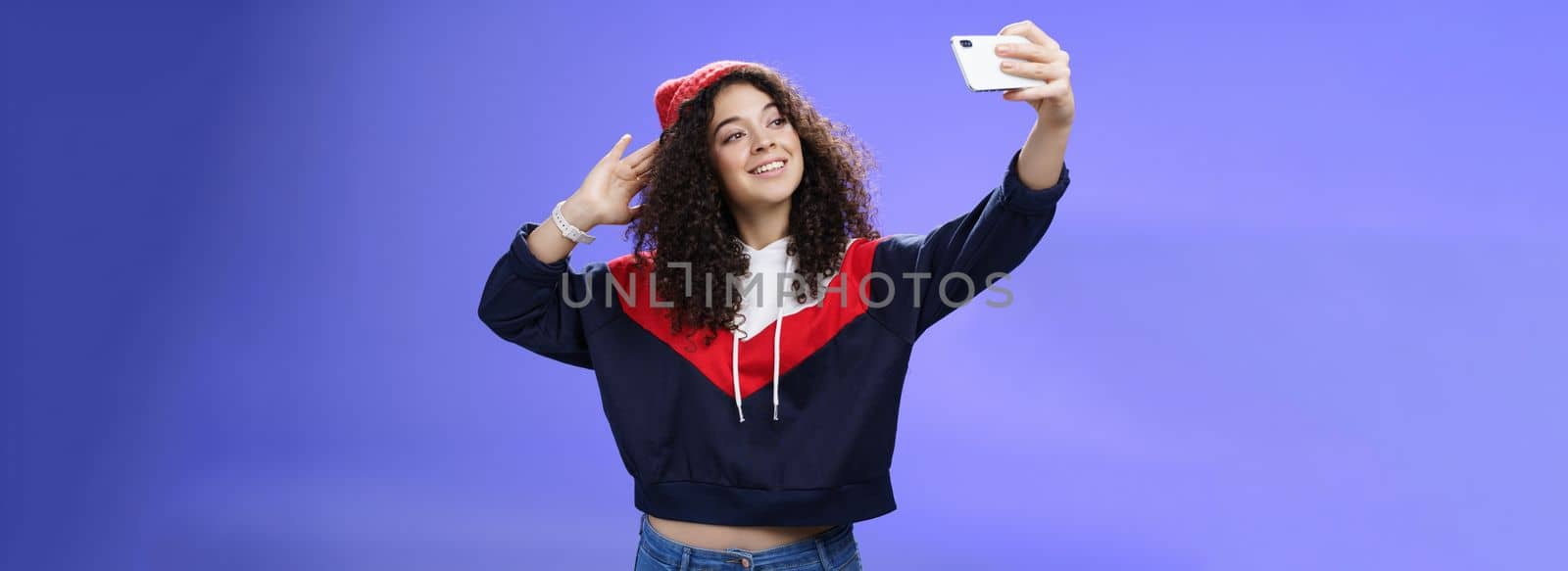 Cute girl making selfie to memorize trip standing in warm winter beanie and sweatshirt posing as pulling hand with smartphone and smiling at device screen to take pretty shot and post internet. Technology and lifestyle concept