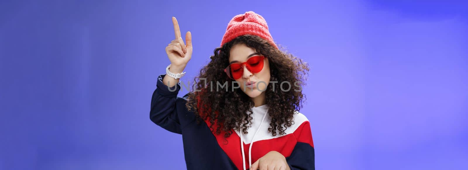 Lifestyle. Cute party girl having fun making disco movements folding lips as enjoying cool song dancing joyfully wearing stylish red sunglasses and warm hat looking down enjoying music at party over blue wall.
